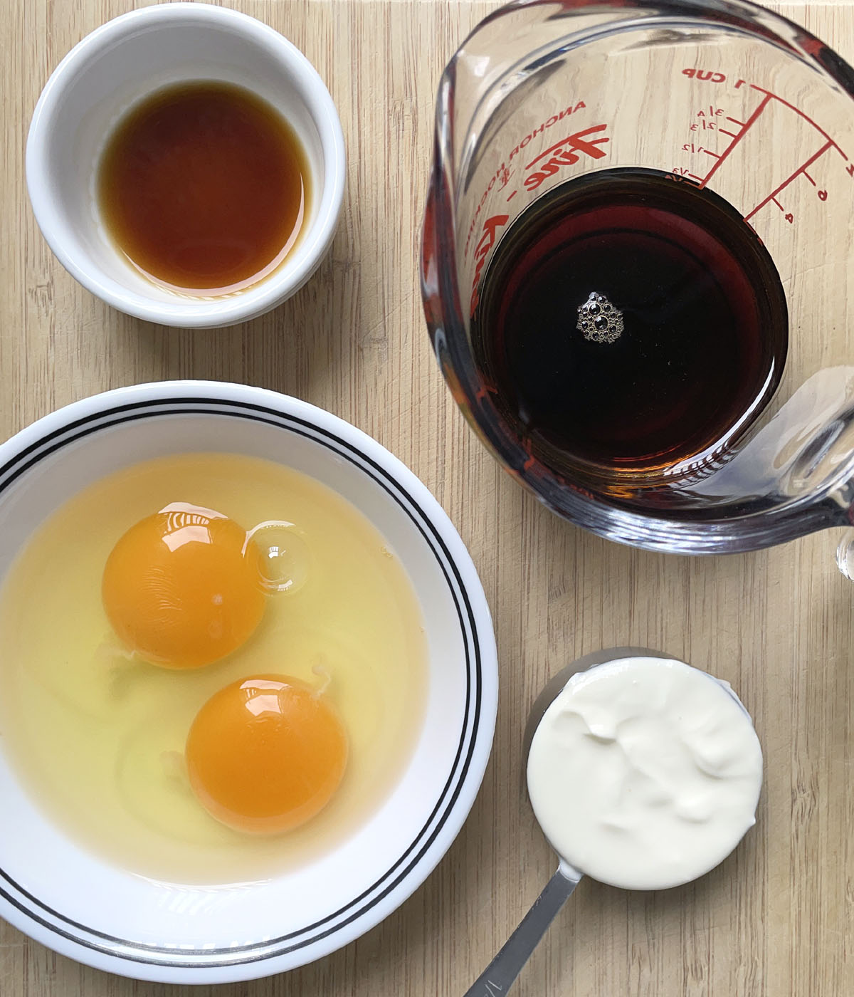 Two raw eggs in a bowl, a measuring cup with white yogurt, a measuring up with brown liquid, a white dish with brown liquid.