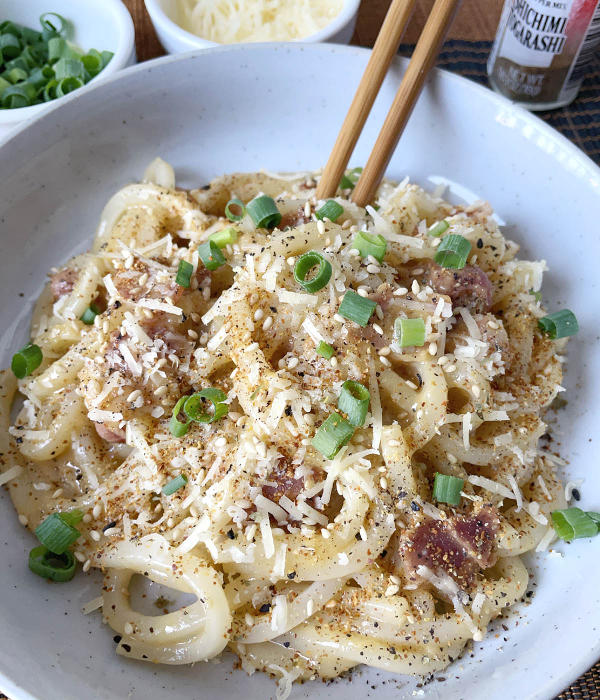 A pair of wooden chopsticks in a dish containing miso udon carbonara topped with chopped green onions and grated cheese.