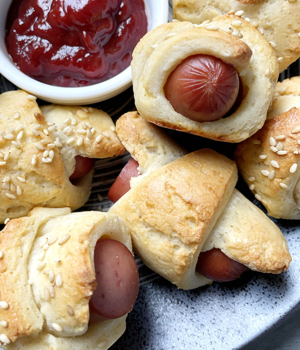 A small white bowl of red ketchup, surrounded by bread wrapped wieners on a plate.