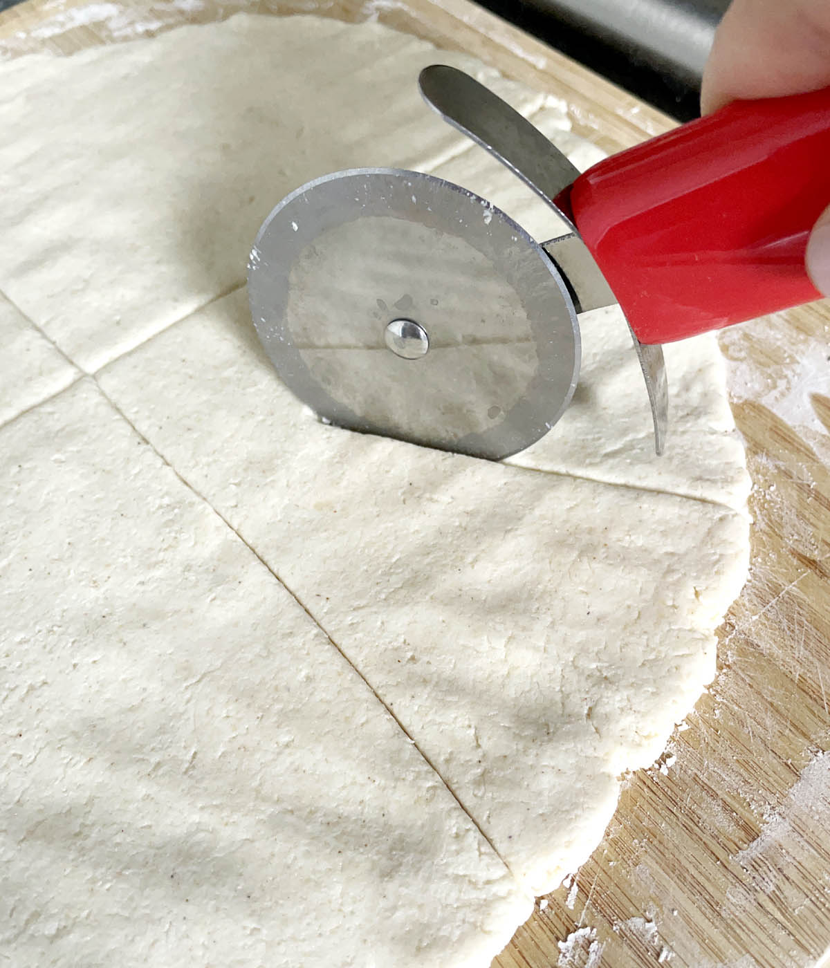 A pizza cutter cutting wedges into dough on wooden cutting board.