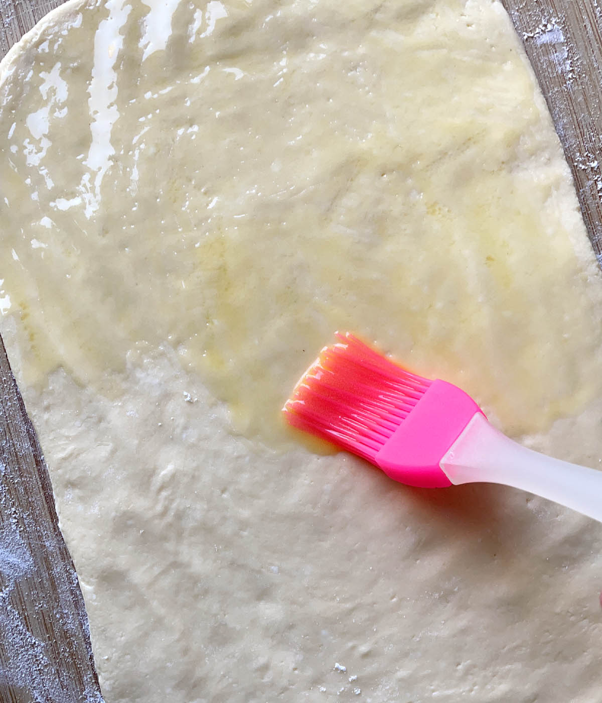 A pink brush brushing melted butter on rectangular shaped dough.