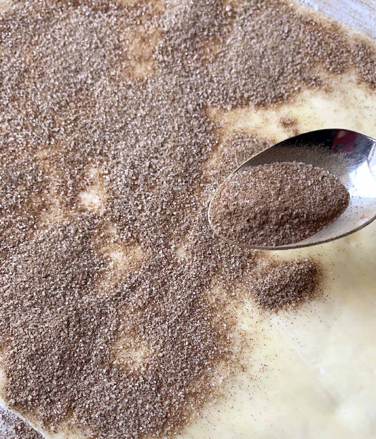 Brown cinnamon and sugar being sprinkled from a spoon onto a dough rectangle.