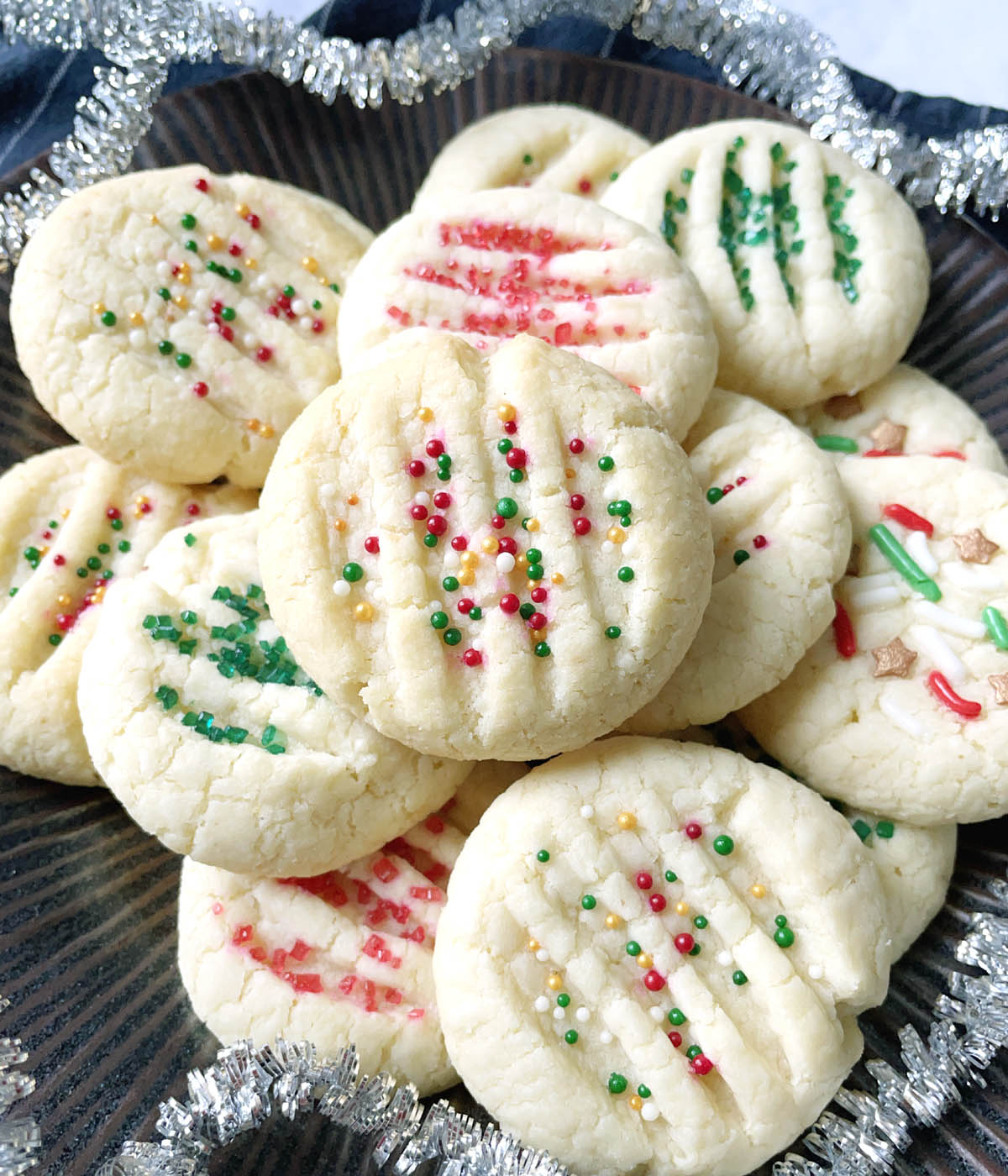 A round brown dish surrounded by silver garland and containing several light colored cookies with red and green and white sprinkles.