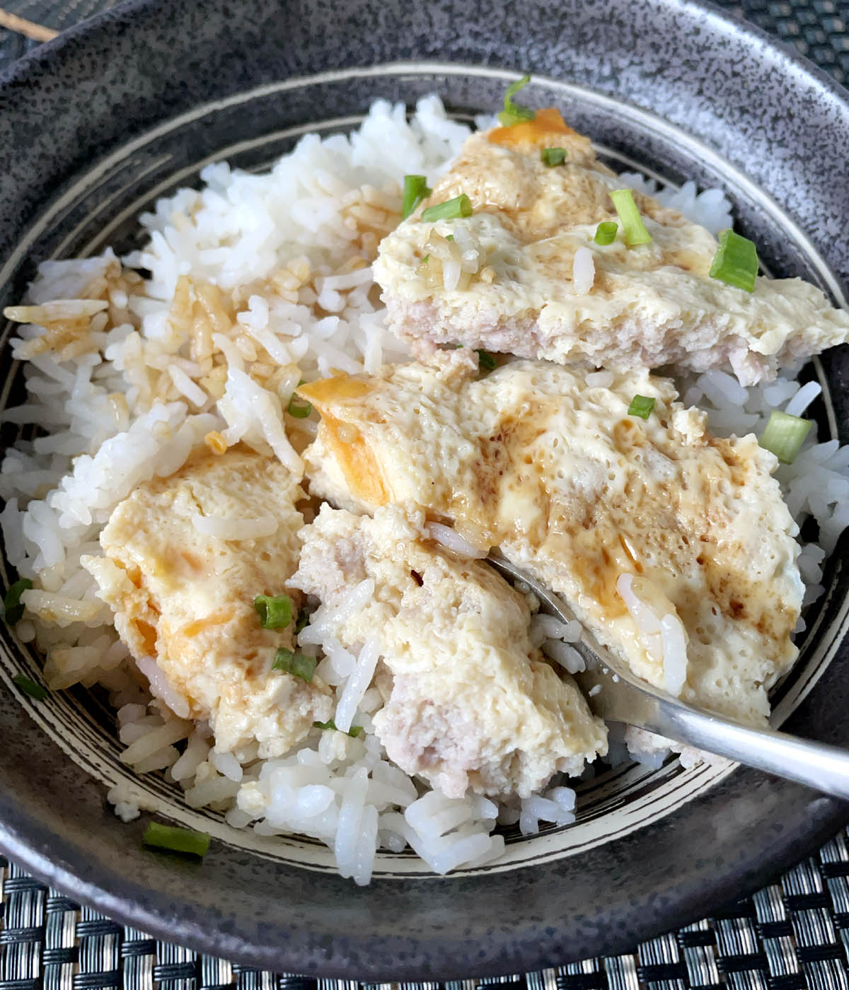 A metal spoon in a dark round bowl containing steamed egg and minced pork patty and white rice.