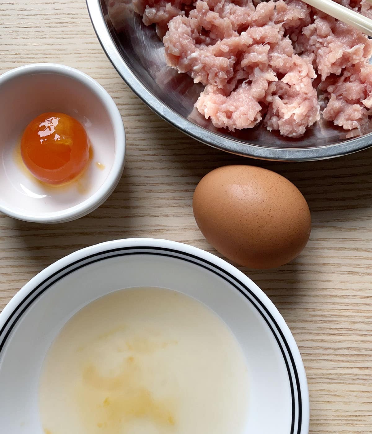 A round metal bowl containing raw pink ground meat, a white dish containing an orange egg yolk, a brown shell egg, and a white bowl containing egg white.