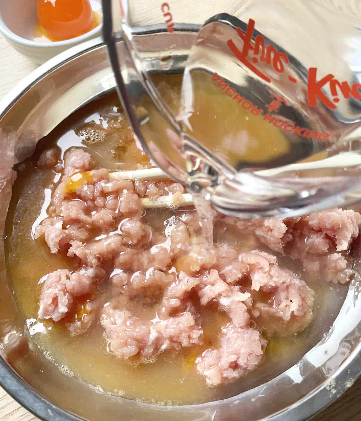 Water being poured into a metal dish containing raw pink ground pork and an egg mixture.