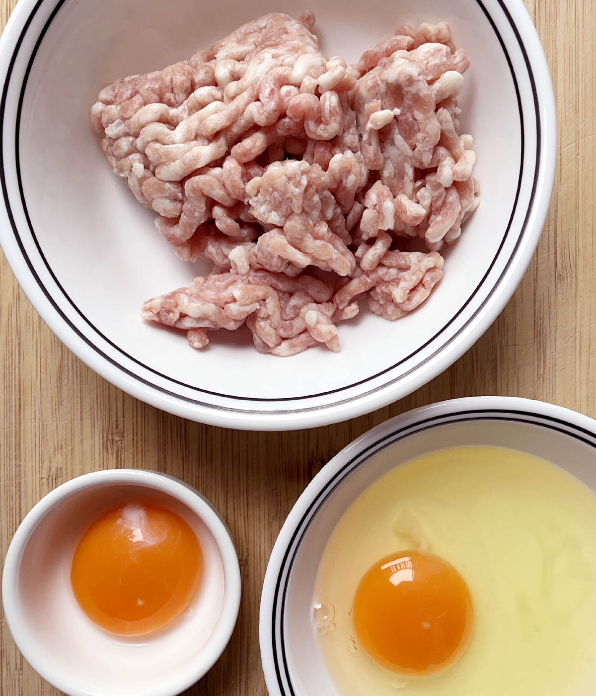 A white bowl containing raw pink ground pork, a white bowl containing an egg yolk, a white bowl containing an egg yolk and egg white.