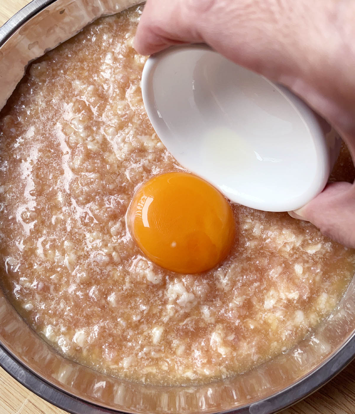 A hand pouring an egg yolk from a white dish onto a raw pink ground mix mixture in a metal dish.