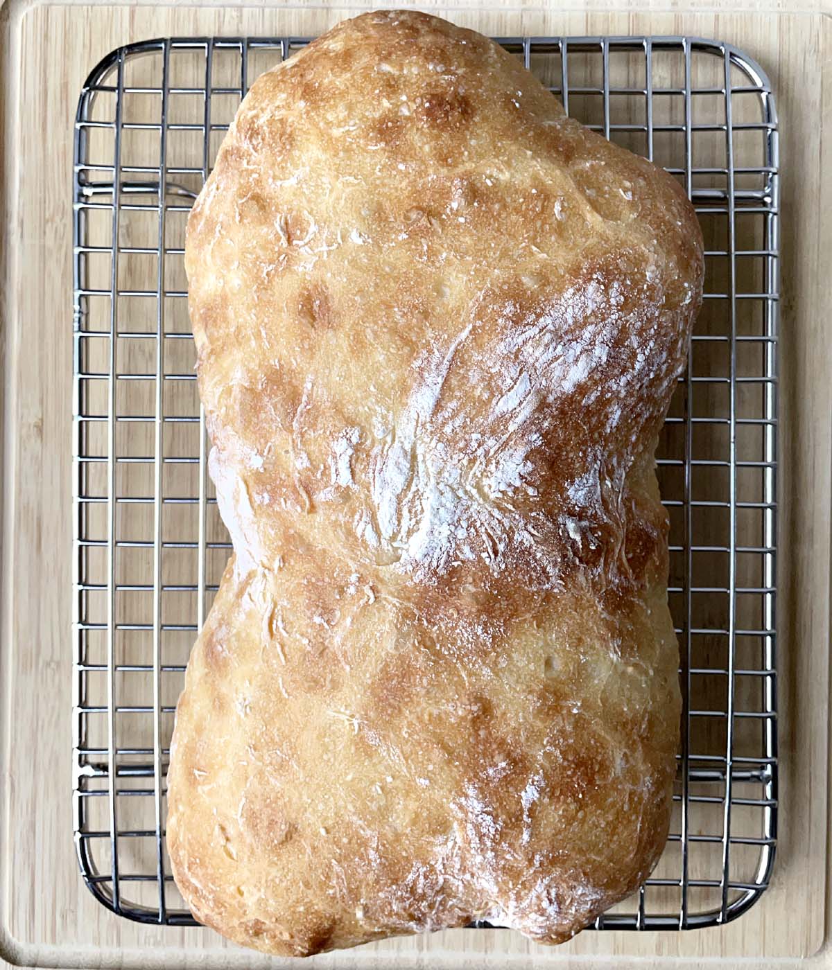A brown baked loaf of bread on a metal rack on a wooden cutting board.