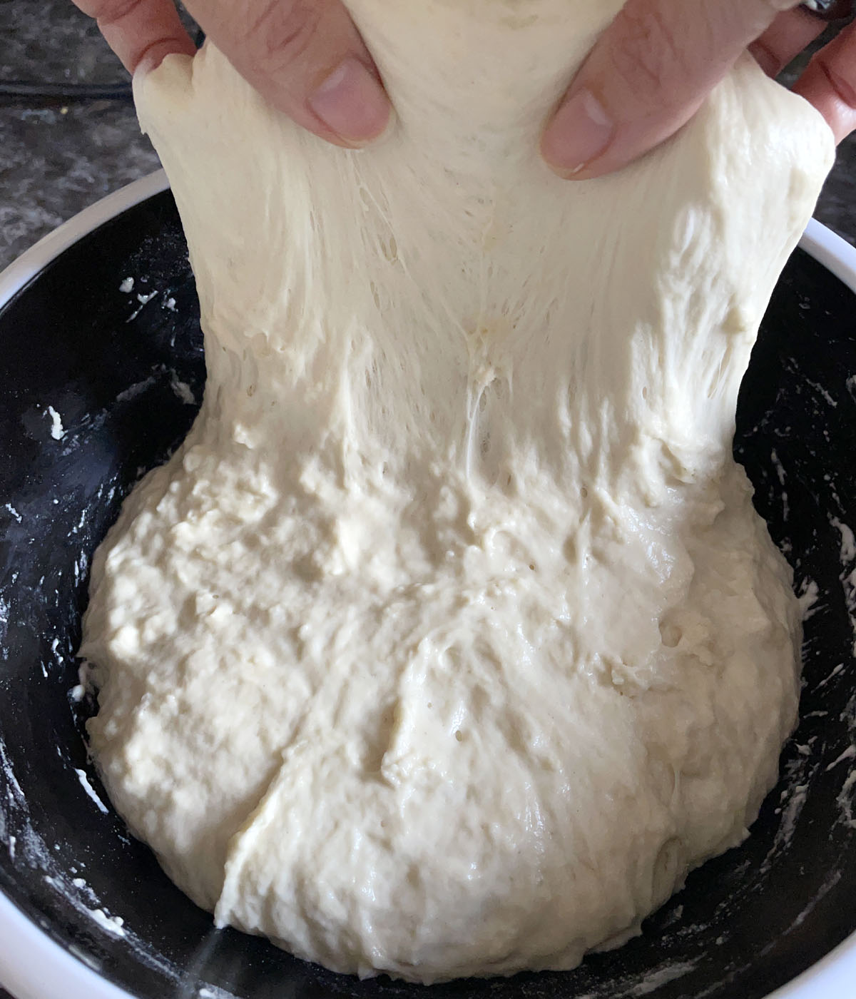 Two hands stretching dough upwards in a bowl.