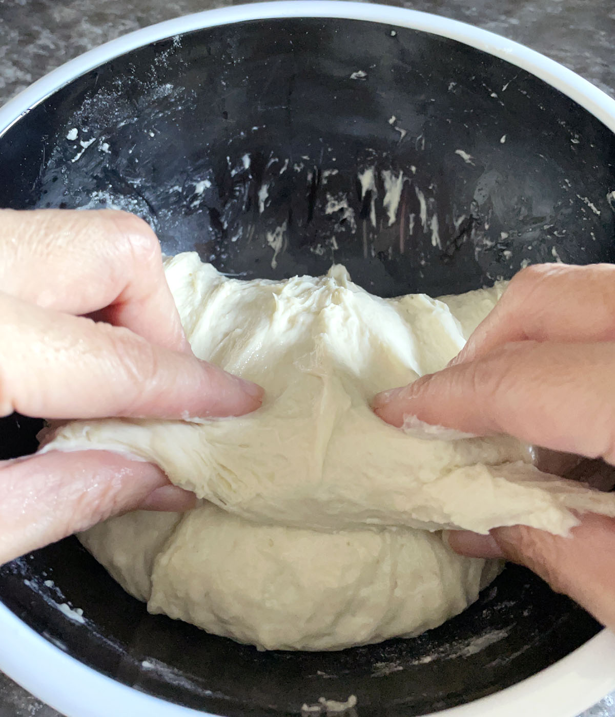 Two hands folding dough over onto itself in a dark bowl.