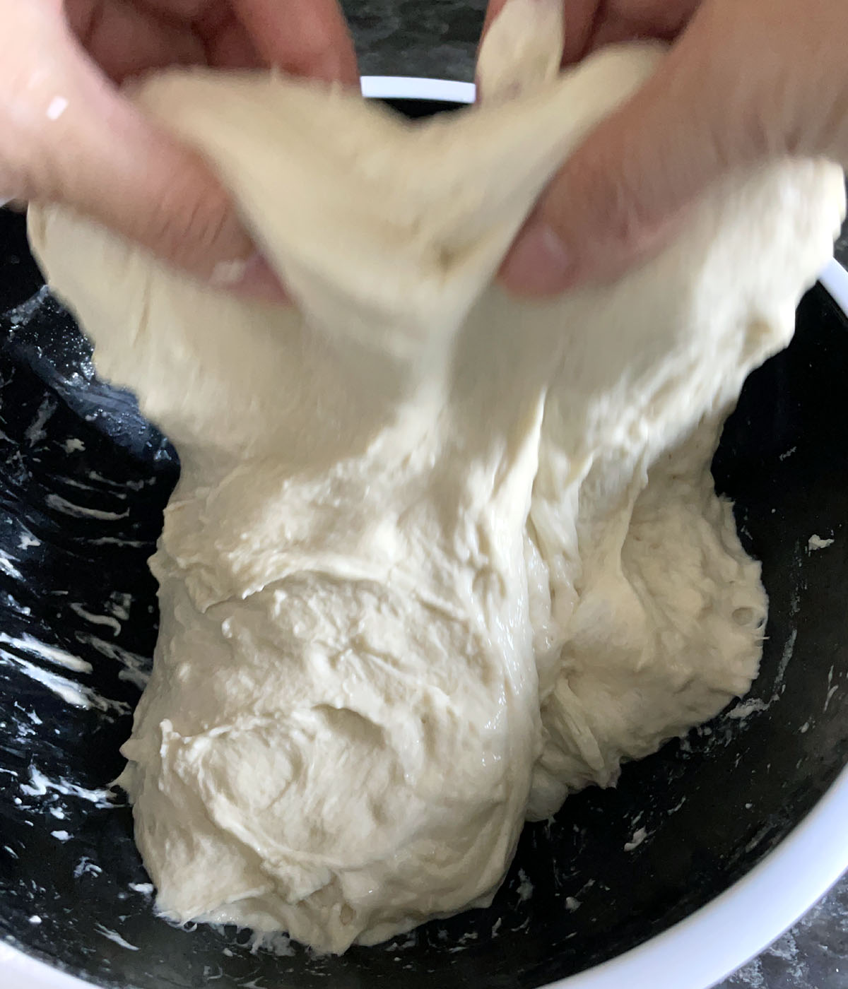 Two hands stretching dough upward over a dark round bowl.