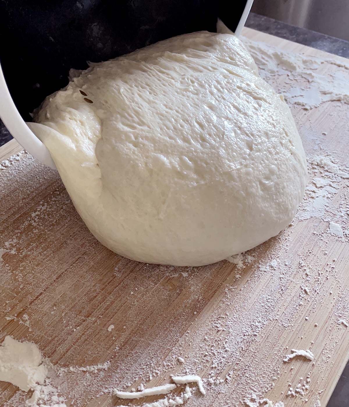 A ball of bread dough being poured out of a bowl and onto a floured wooden cutting board.