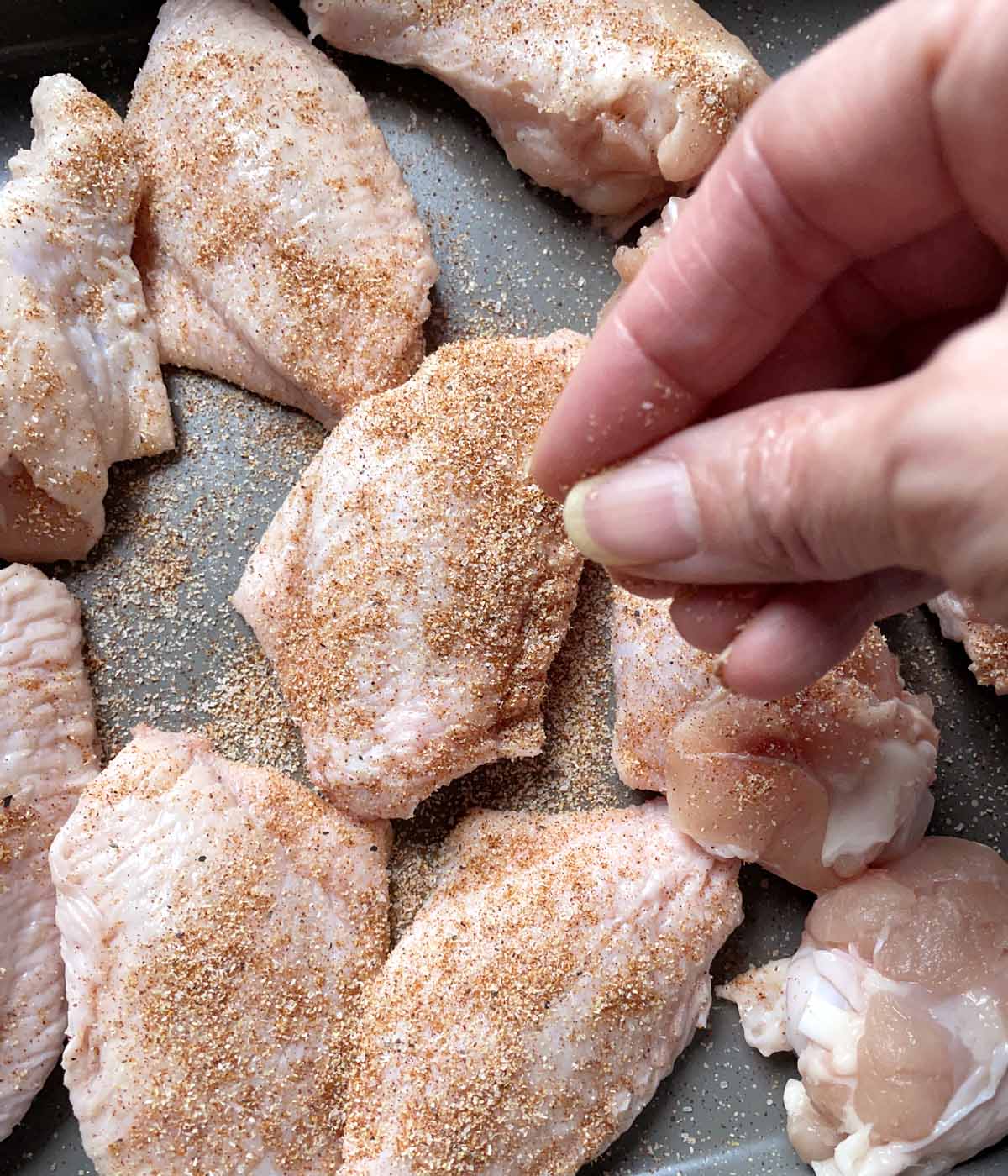 A hand sprinkling brown spices over raw chicken wings in a pan.