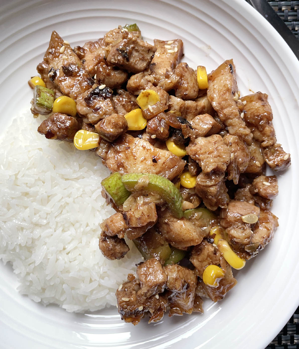 A round white bowl containing cooked white rice and black bean garlic pork belly, green zucchini, and yellow corn.