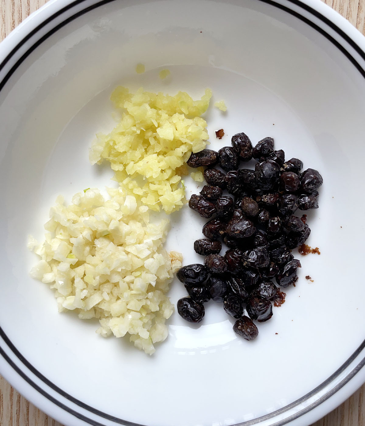 A round white bowl containing black beans, minced ginger, and minced garlic.