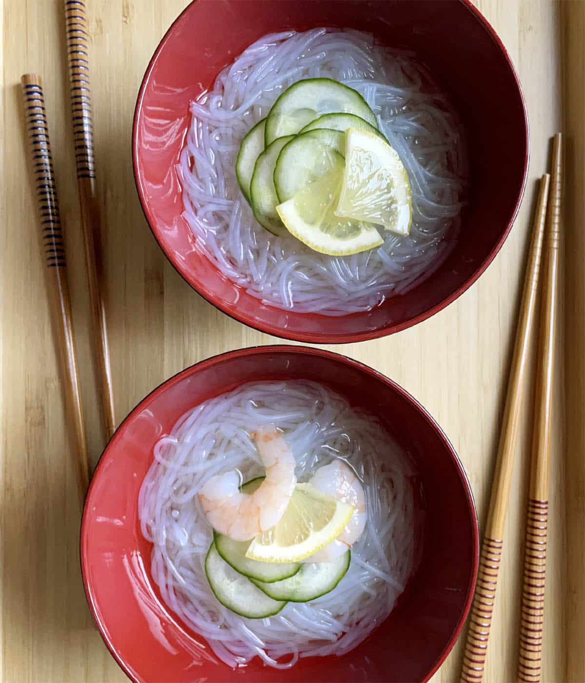 Two pairs of wooden chopsticks next to two red bowls containing white noodles, cucumber slices, cooked shrimp, and lemon slice wedges.