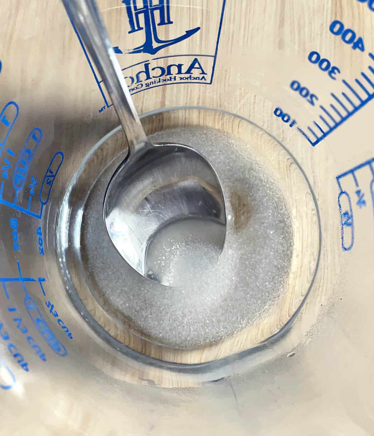 A glass measuring cup containing a clear liquid, sugar, and a spoon.