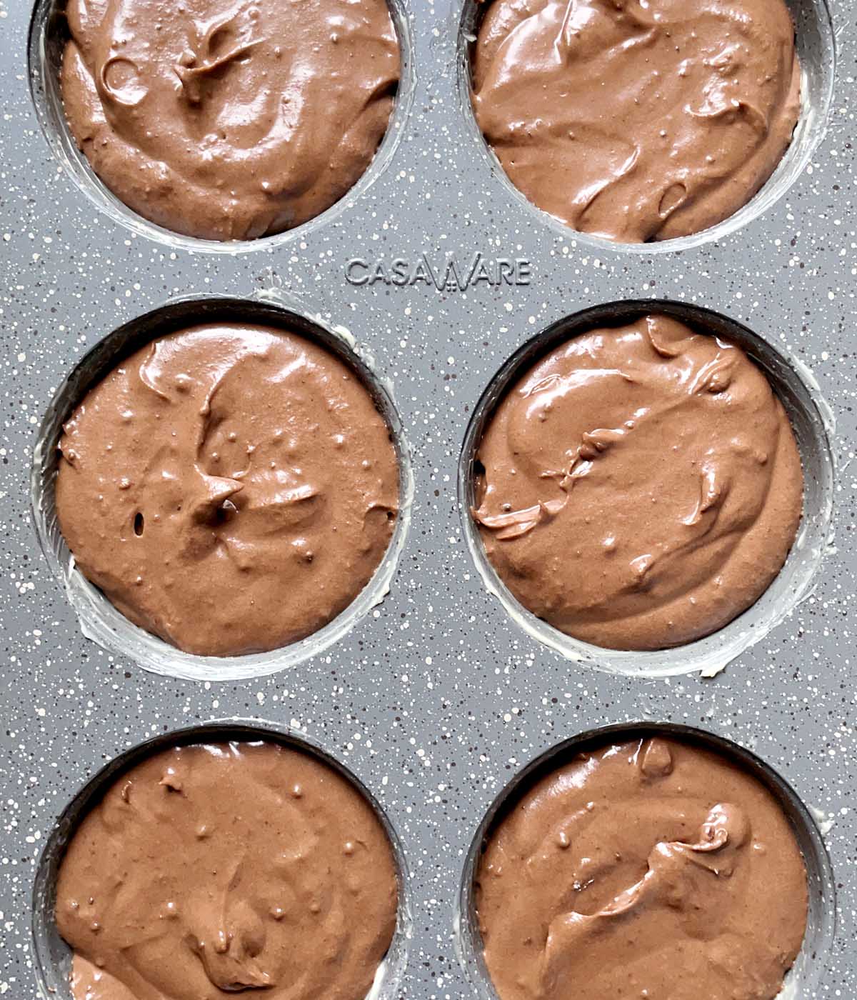 Six muffin cups in a grey metal muffin tin containing brown batter.