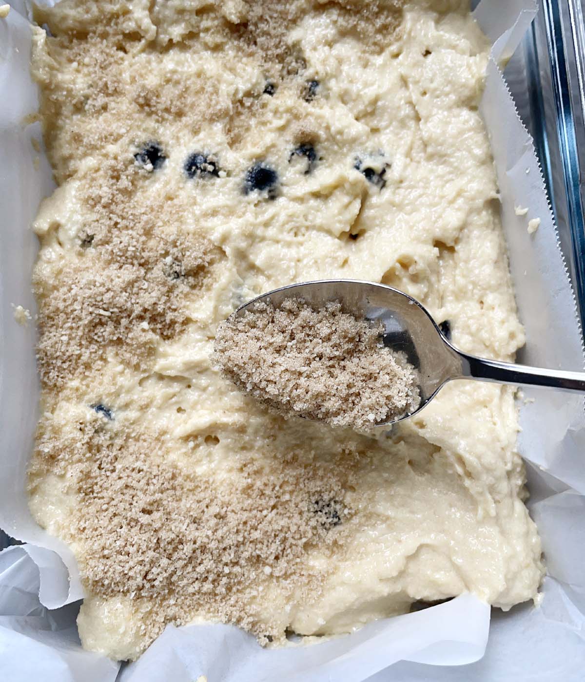 A spoon sprinkling a brown crumbly flour over cake batter in a rectangular dish.