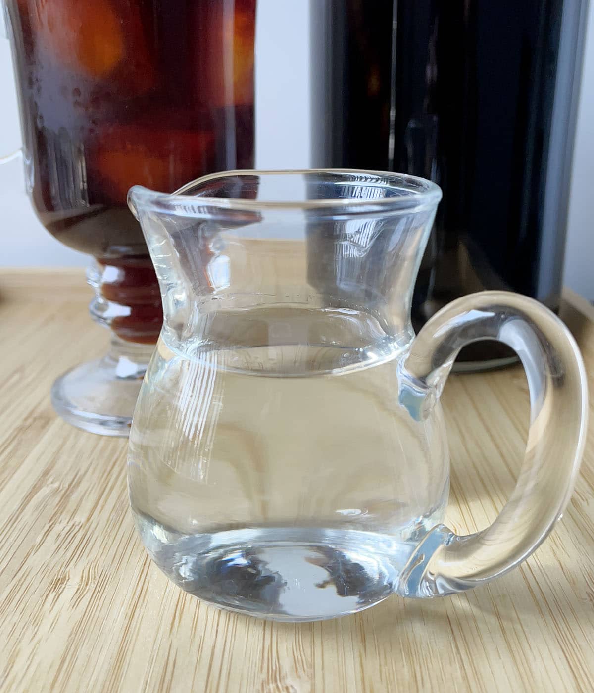 A small glass pitcher of clear simple syrup in front of a glass of iced coffee and a bottle of brown liquid.