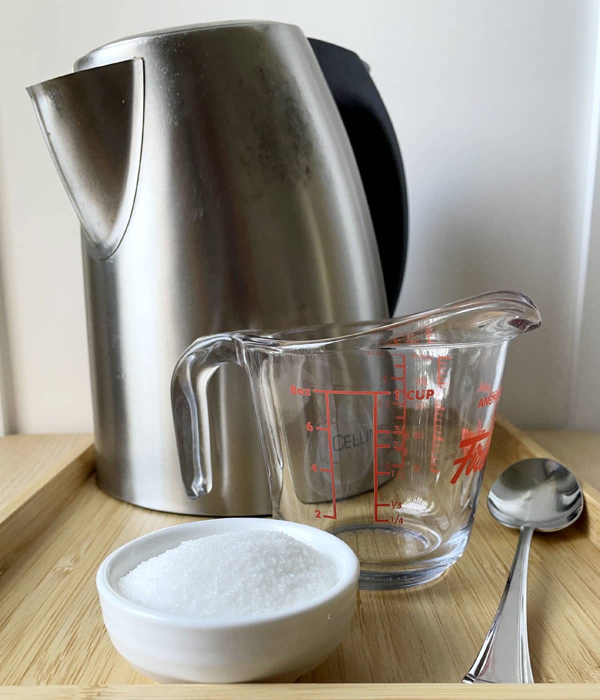 A metal kettle, an empty glass measuring cup, a spoon, and a white round dish containing sugar.