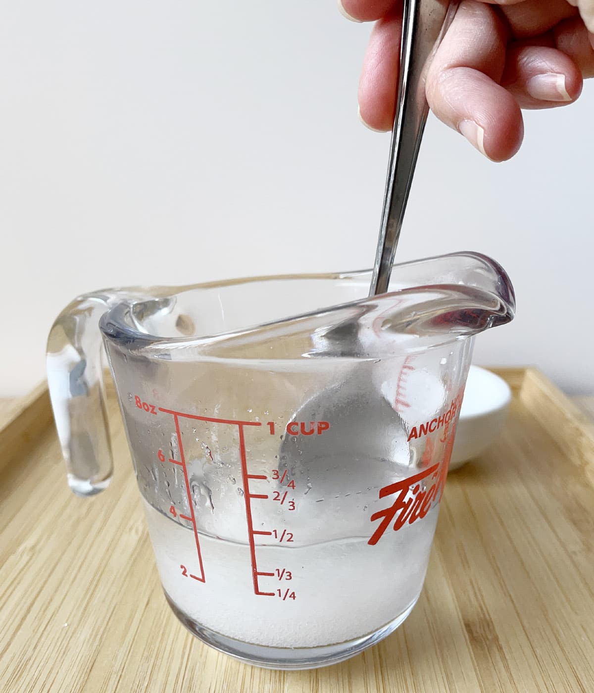 A hand holding a spoon, stirring white sugar in a water in a glass measuring cup.