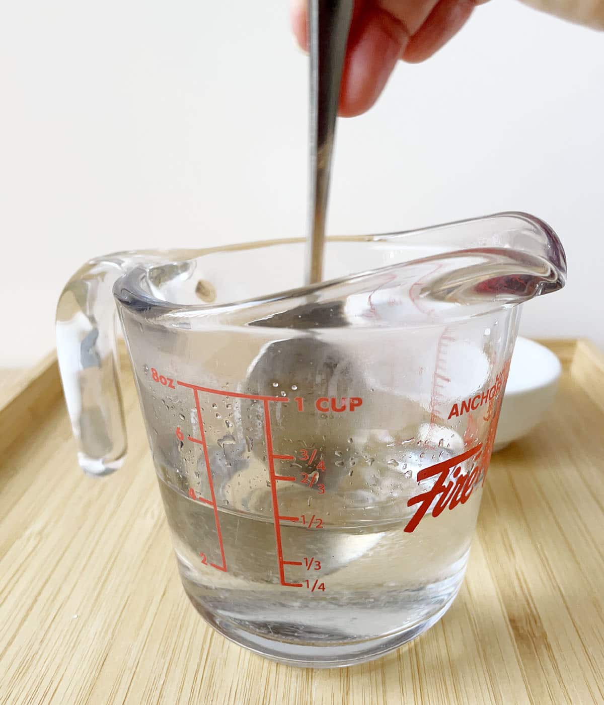 A hand holding a spoon stirring clear liquid in a glass measuring cup.