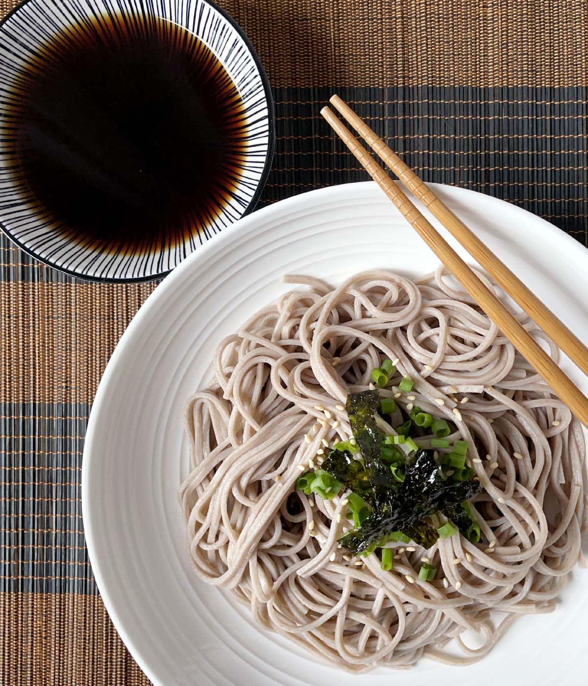 A pair of wooden chopsticks resting on a white plate containing soba noodles. A small black and white bowl containing dark liquid.