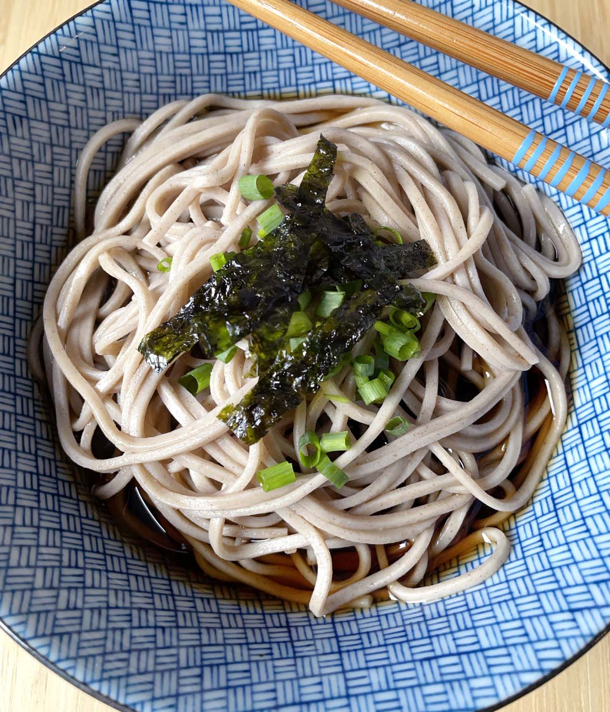 A pair of wooden chopsticks on a blue bowl containing light brown soba noodles and brown sauce, topped with chopped green onions and seaweed strips.