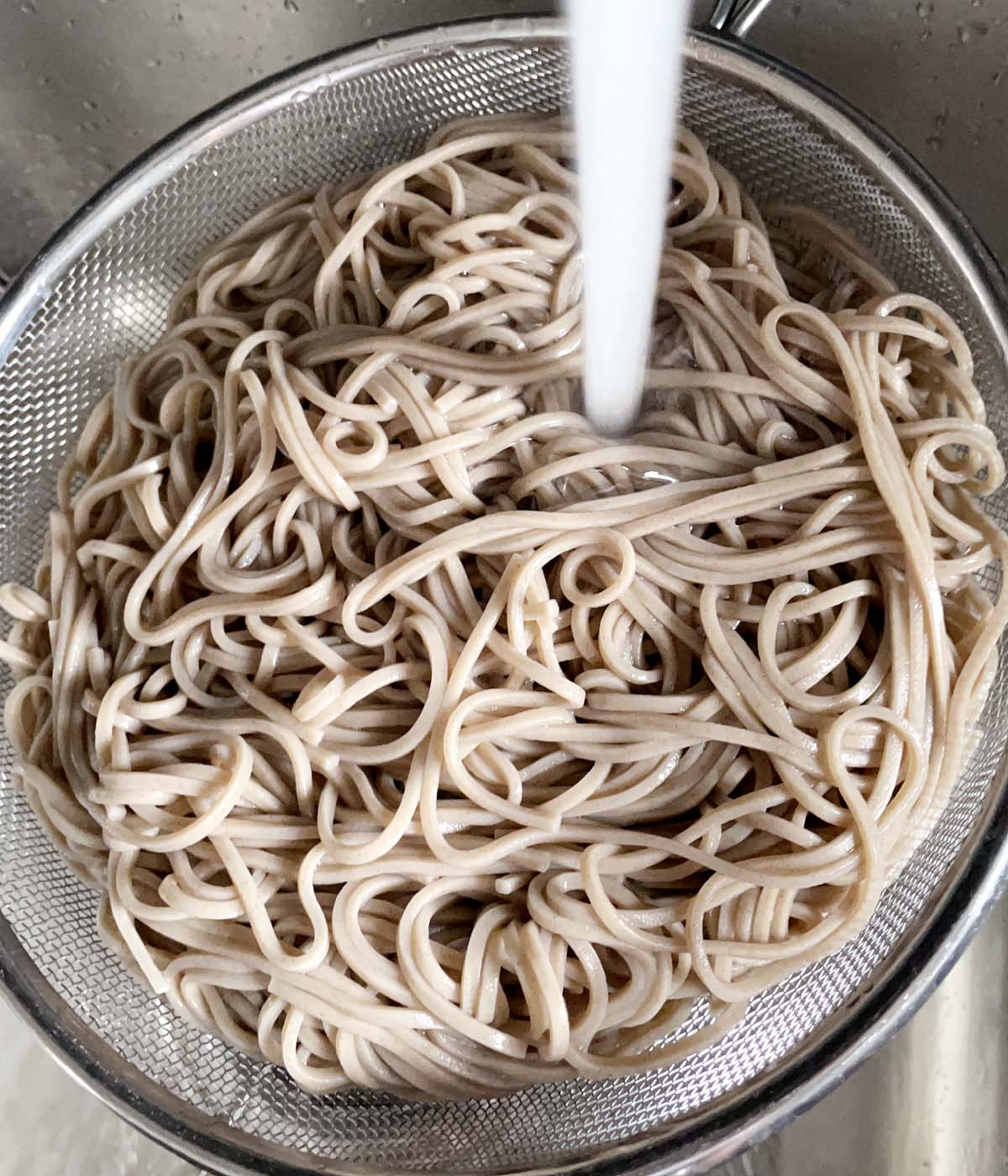 Water rinsing cooked light brown noodles in a metal colander.