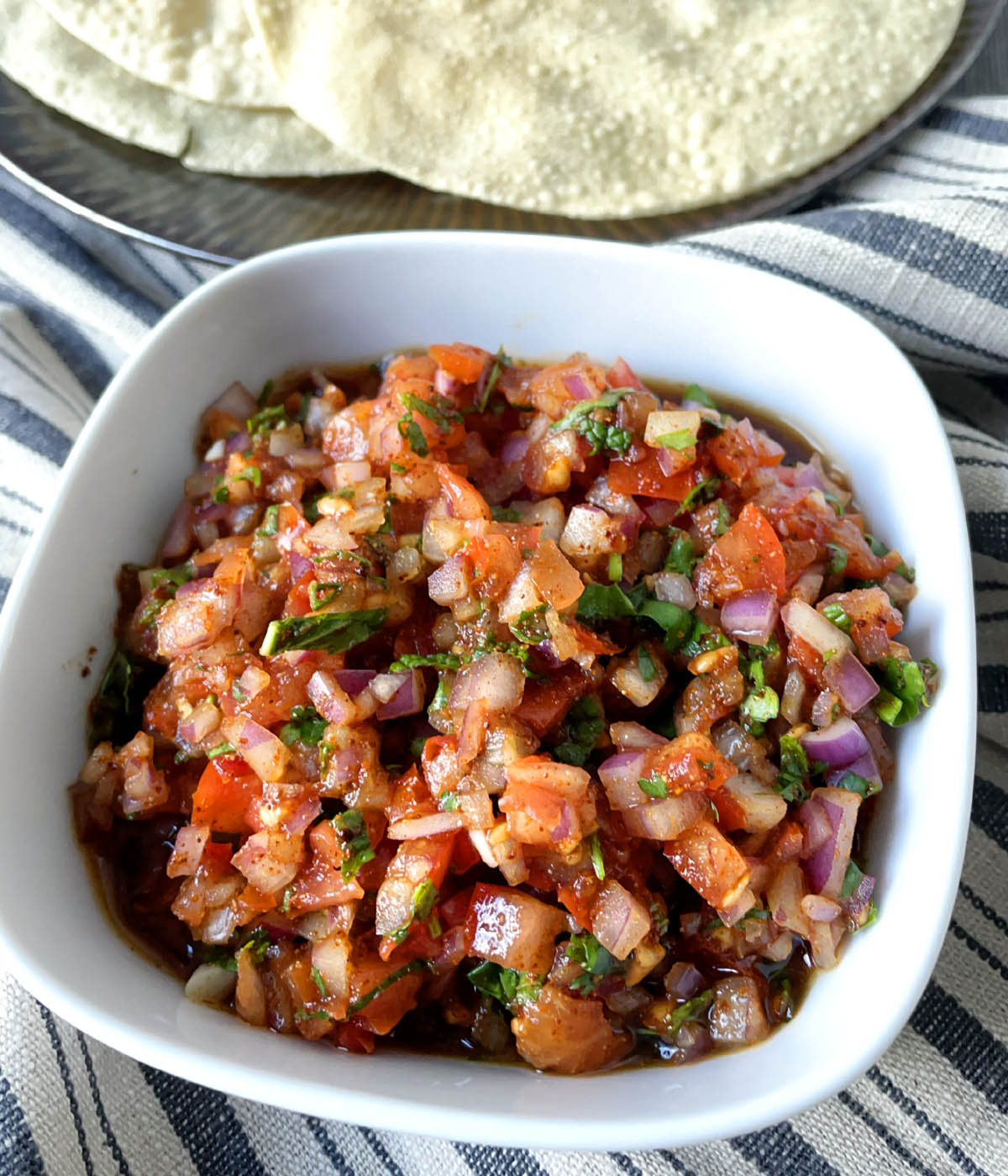 A white square dish containing finely diced red tomatoes, red onions, and chopped green herbs.