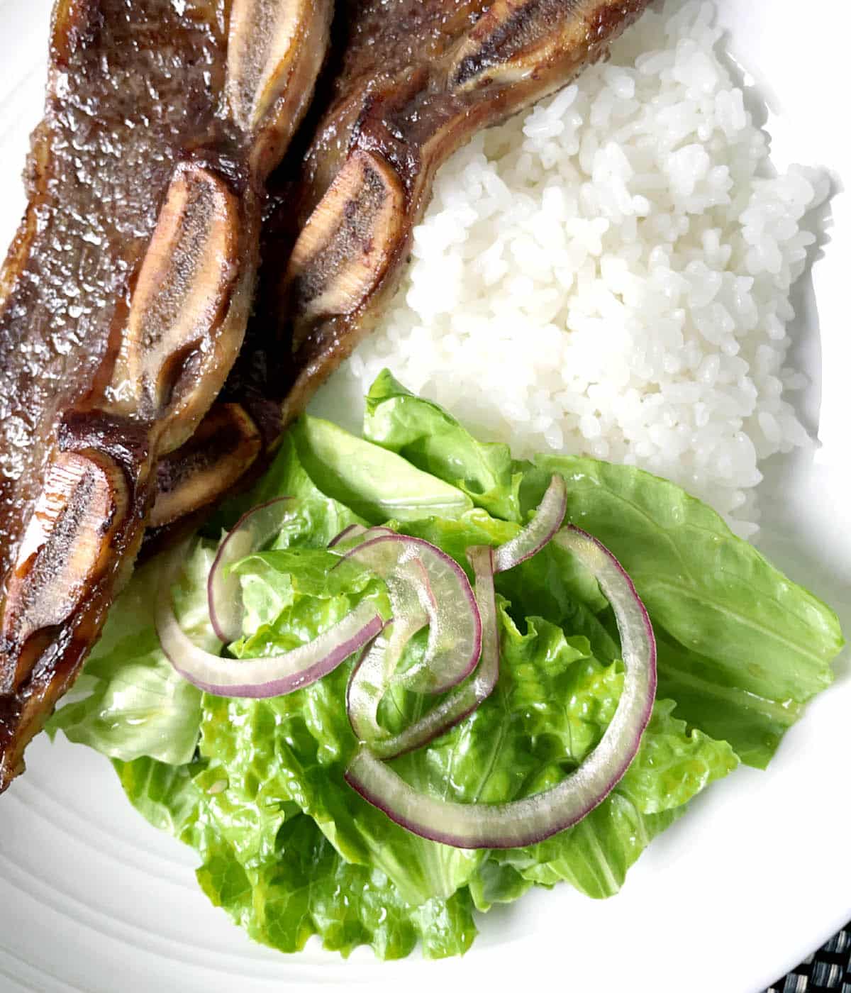 A white plate containing white rice, cooked beef ribs, green lettuce leaves and thinly sliced red onion.