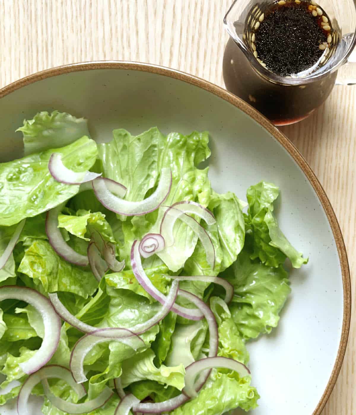 A small glass pitcher containing brown liquid, next to a round plate with a brown edge containing green lettuce leaves and thinly sliced red onion.