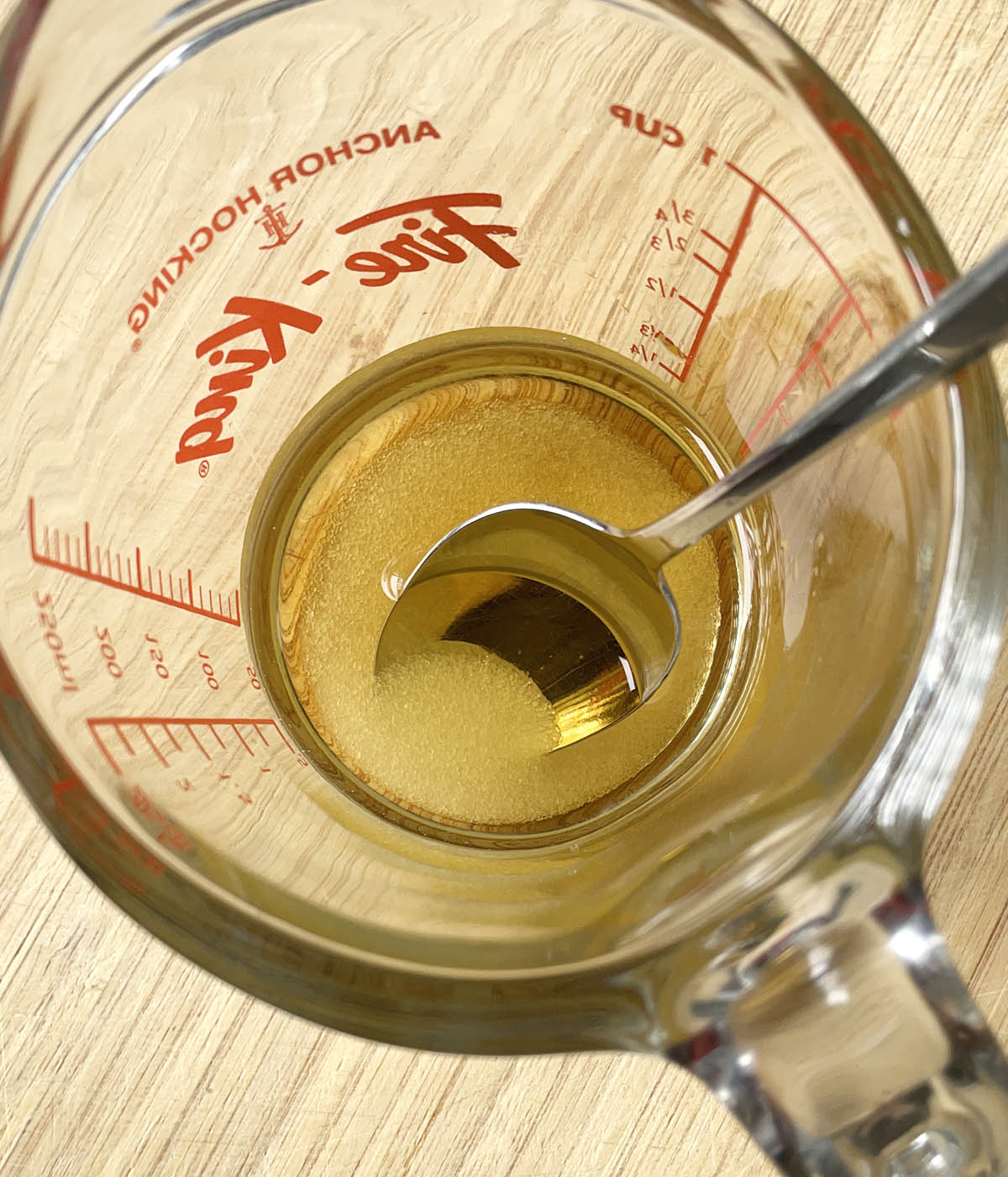 A glass measuring cup containing a clear yellow liquid, white sugar, and spoon.