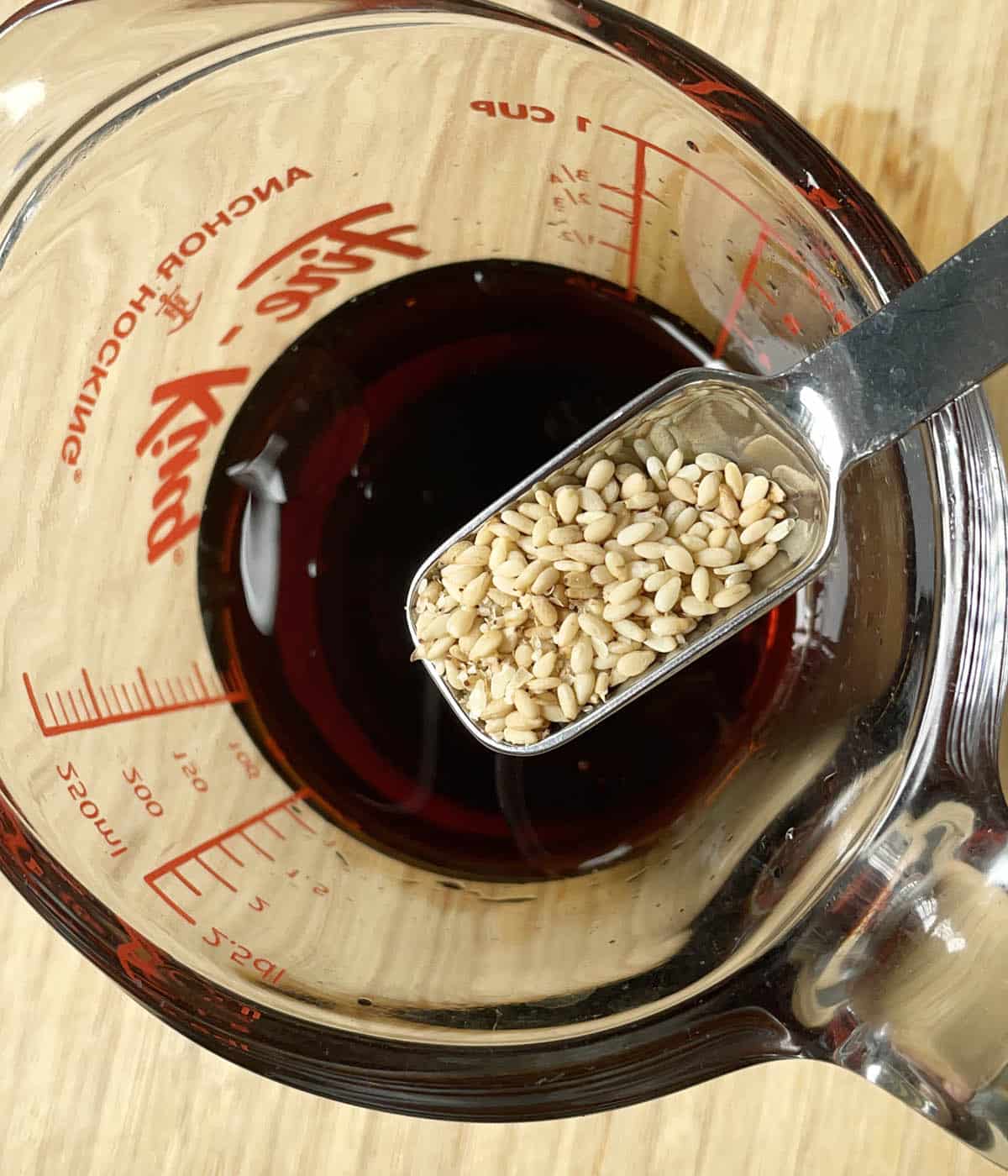 A metal spoon containing sesame seeds over a glass measuring cup containing brown liquid.
