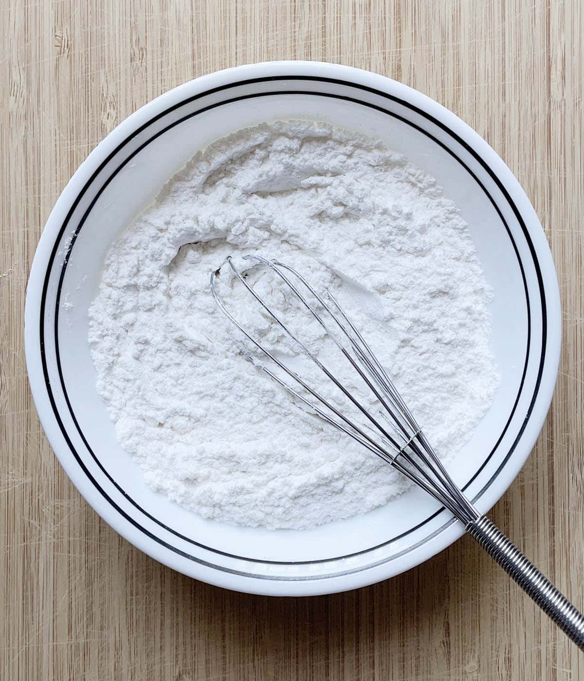 A metal whisk in a round white bowl containing white flour.