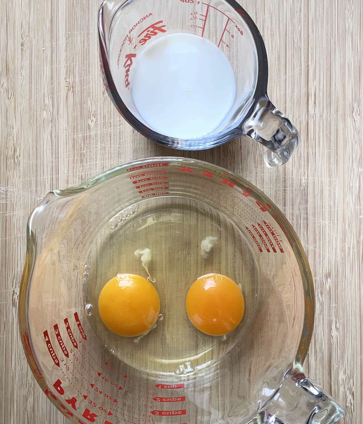 A large glass measuring cup containing two yellow egg yolks and whites, and a small glass measuring cup containing white milk.