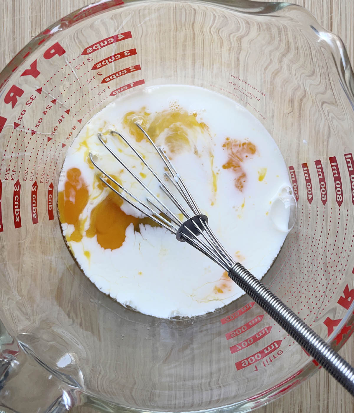 A metal whisk resting in a large glass measuring cup containing white milk and egg yolks.