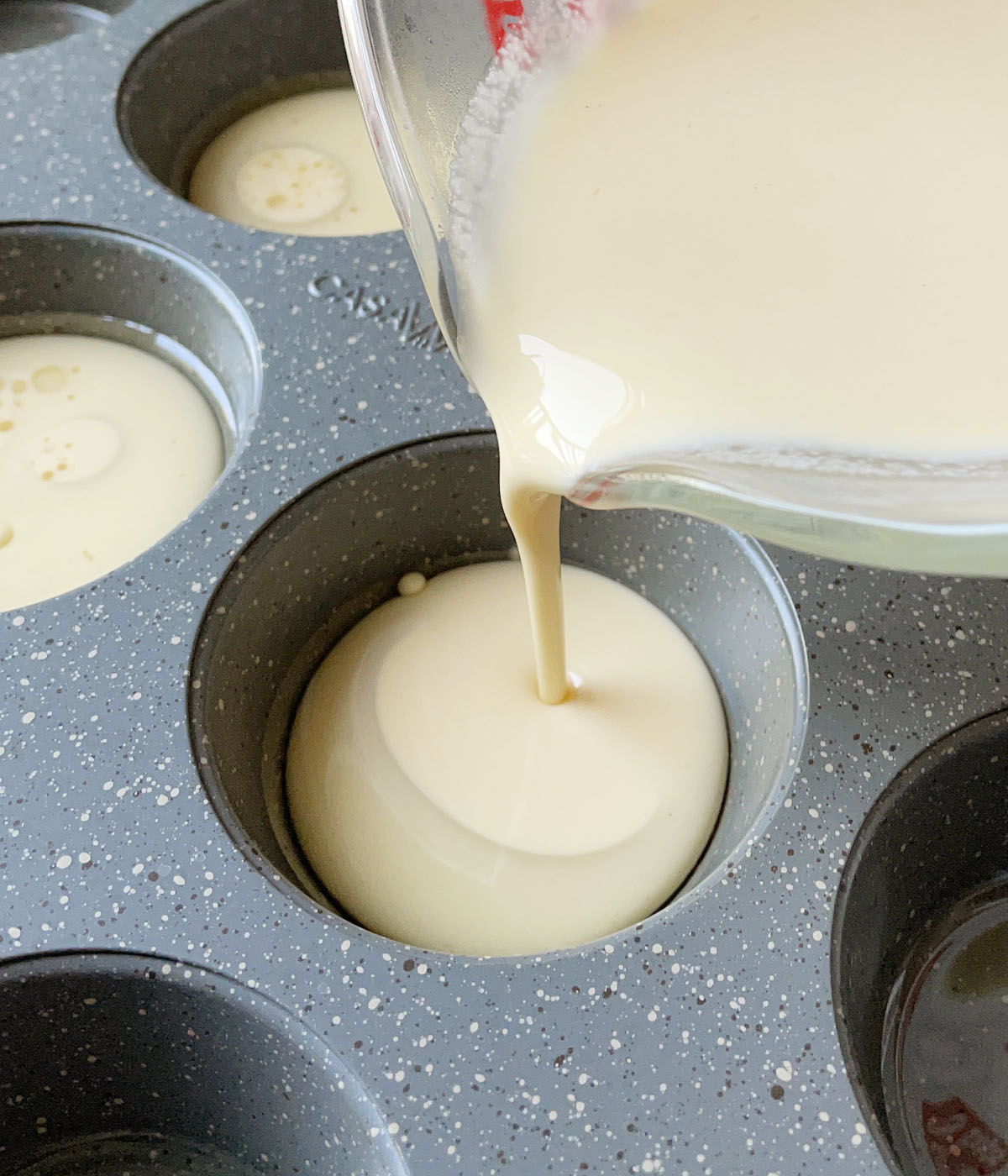 A light yellow liquid batter being poured into a metal muffin tin.