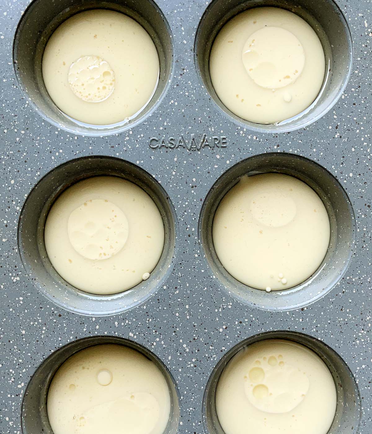 A grey speckled metal muffin tin containing light yellow batter in the cups.