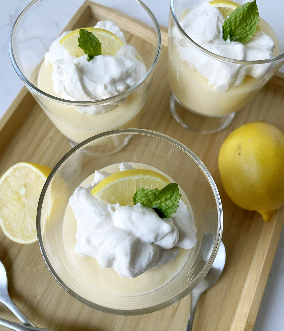 Three glass dishes containing yellow pudding, whipped cream, a lemon wedge, and spearmint leaf.