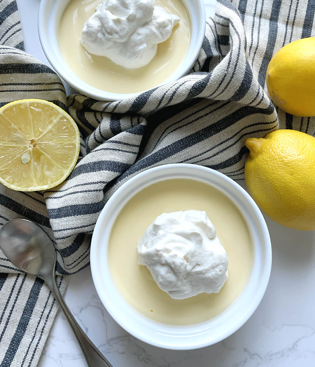 A striped towel and lemons next to two white round dishes containing lemon pudding and whipped cream.