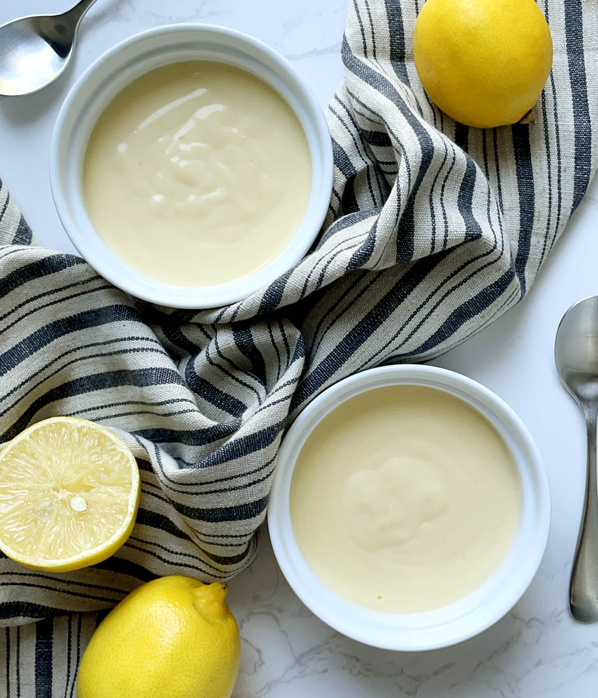 Two round white dishes containing lemon pudding, two spoons, a whole lemon, and a cut lemon.