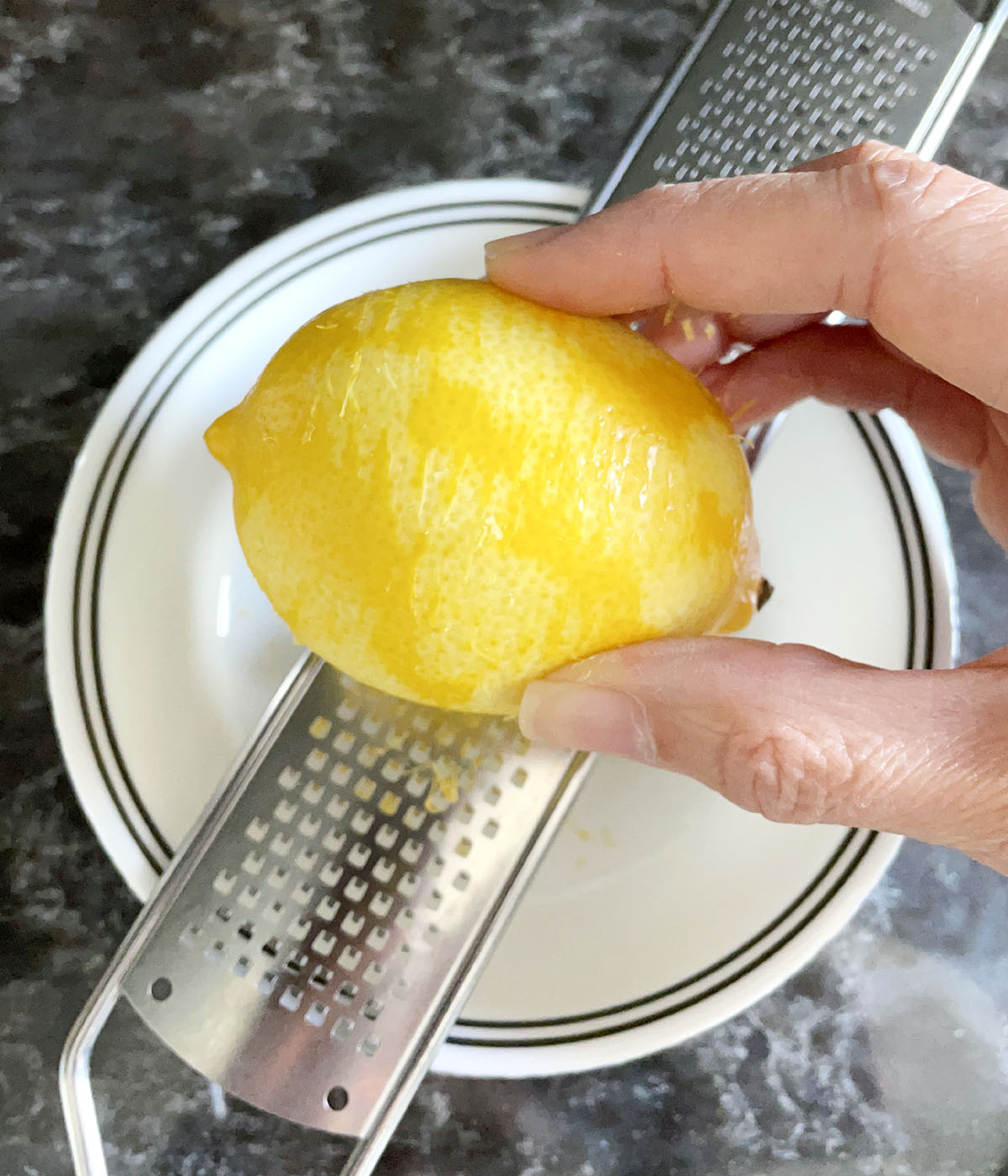 A hand holding a yellow lemon over a microplane.