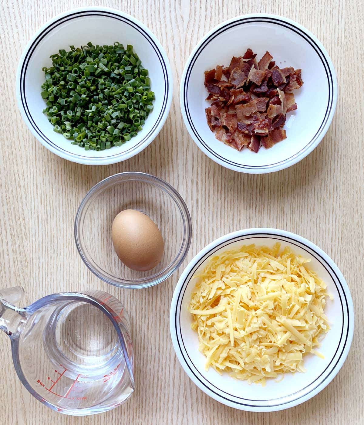 Bowls containing chopped green onions, crumbled cooked bacon, grated cheddar cheese, an egg, and a measuring cup with water.