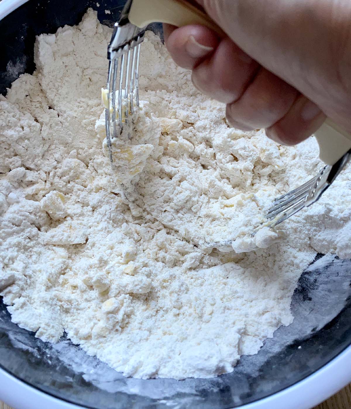 A hand holding a pastry cutter, cutting chunks of butter into dry flour ingredients in a black bowl.