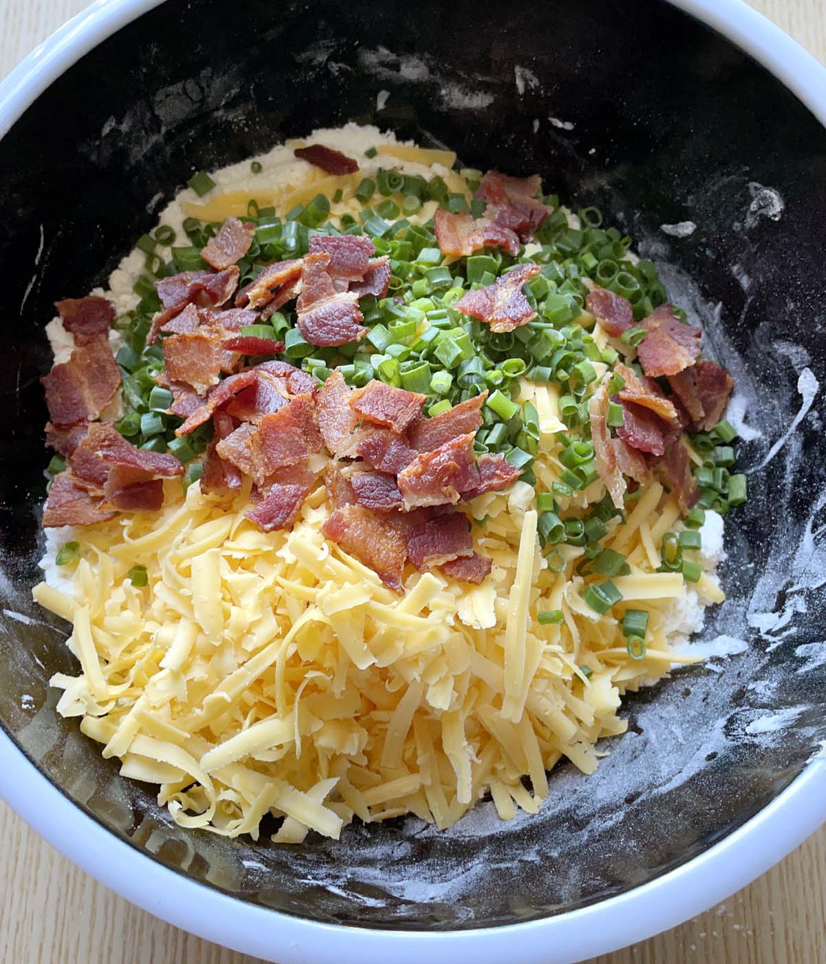 Orange grated cheese, crumbled cooked bacon, and chopped green onions in a round black bowl.