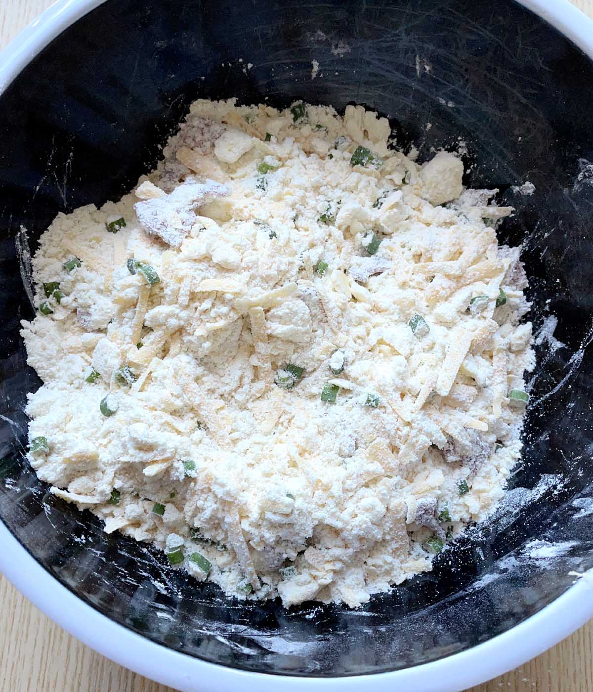 Grated cheese, crumbled bacon, and green onions coated with a flour mixture in a bowl.